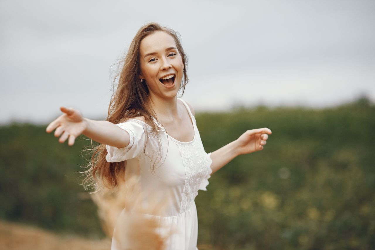 Pomoc i wsparcie Smiling woman in a white dress enjoying nature with open arms.