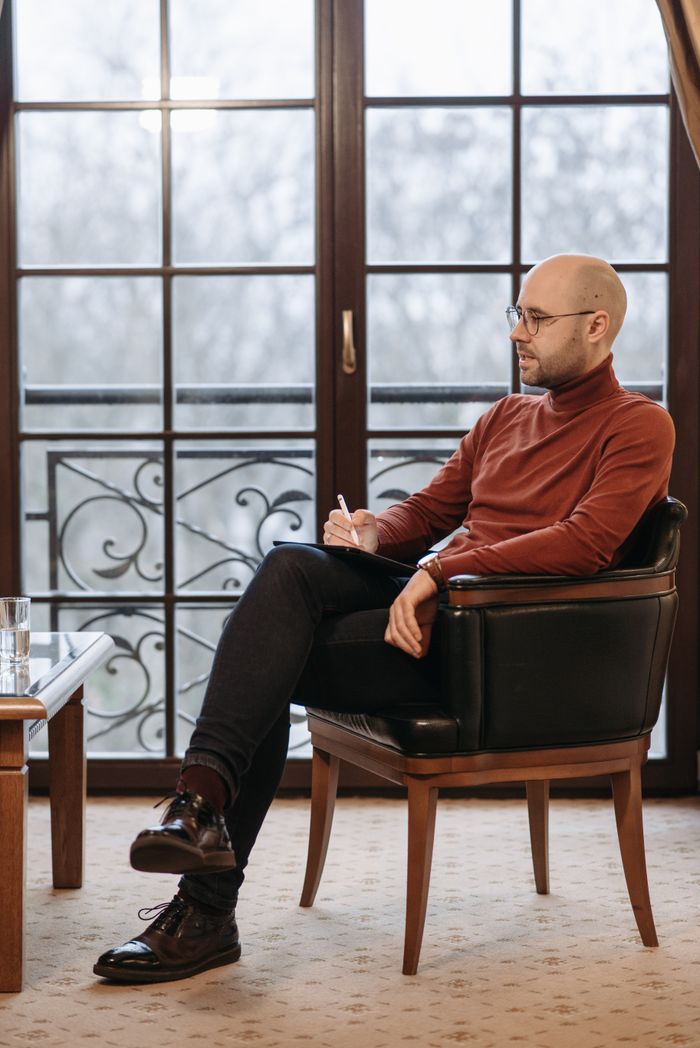 O Fundacji Bald man in sweater sitting with notes near large glass door, looking thoughtful.